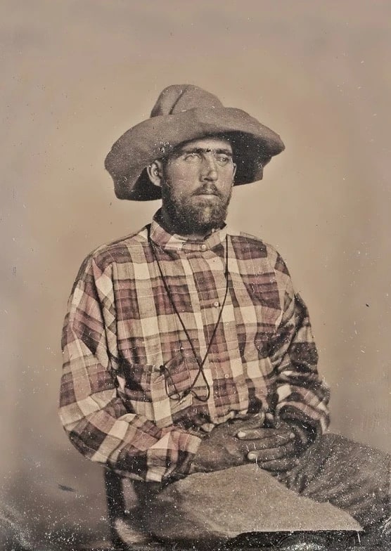 A hand-tinted tintype of a bearded man in a plaid shirt and wide-brimmed felt hat, seated with his hands folded, long identified as Texas Ranger Rufe Perry.