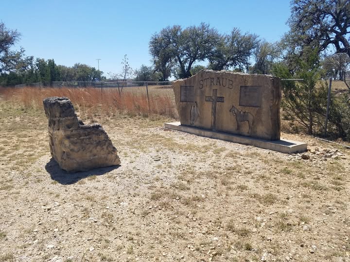 The Straub family monument, a rough-hewn limestone slab carved with a cross, a saddled horse, and a cowboy, with a stone chair beside it and the fence line and pasture beyond.