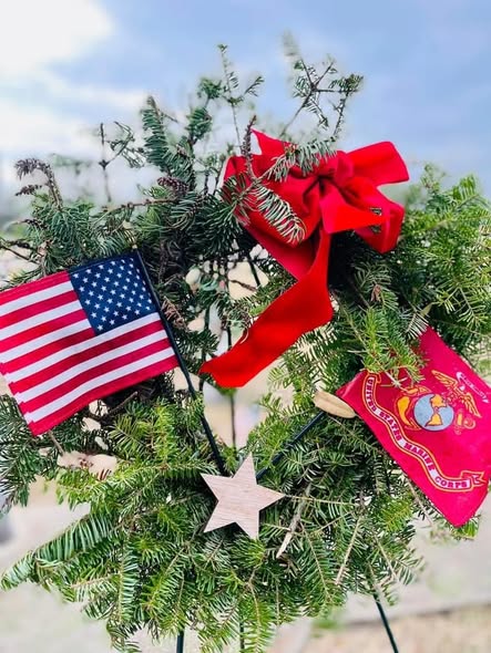 A balsam wreath with a red bow, a small American flag, a Marine Corps flag, and a wooden star.